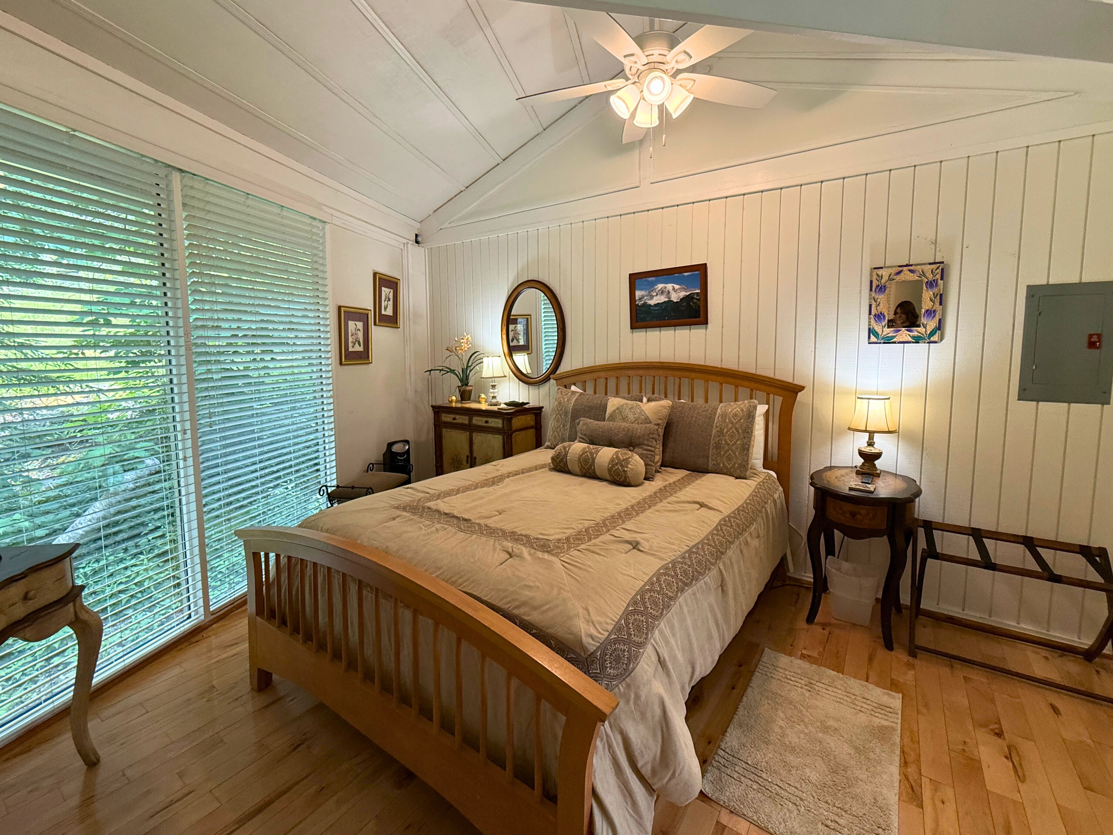 A bedroom with a wooden bed, a ceiling fan, a nightstand with a lamp, a dresser, and a window with blinds.