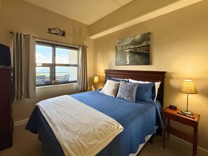 A hotel room with a bed, a window with curtains, a painting on the wall, and a bedside table with a lamp and a clock. - Escape to the beauty of Sugar Mountain in this comfortable 2-bedroom condo (Sugar Mountain)