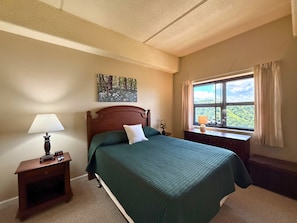 A bedroom with a bed, a nightstand with a lamp, a window with curtains, and a painting on the wall. - Escape to the beauty of Sugar Mountain in this comfortable 2-bedroom condo (Sugar Mountain)