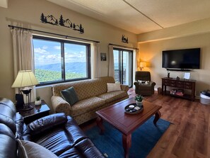 Un salon comprenant un canapé, une table basse, un téléviseur et une vue sur les montagnes grâce à de grandes fenêtres.