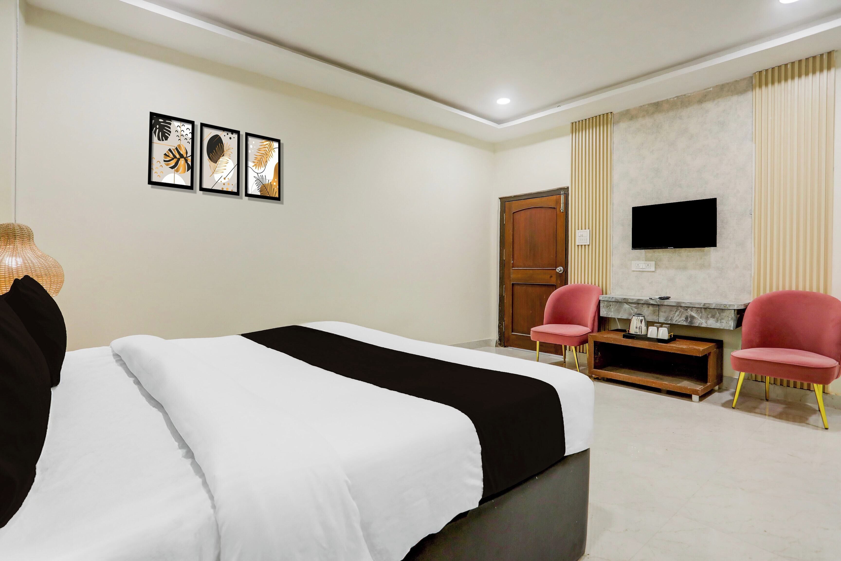 A hotel room with a bed, two chairs, a TV, and artwork on the wall.