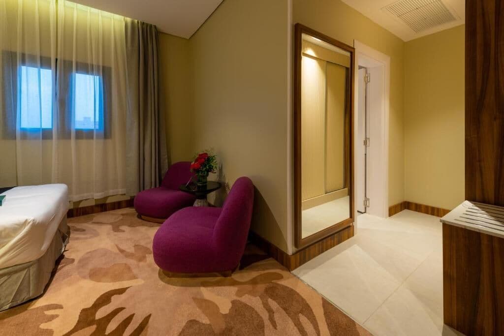 Habitación de hotel con cama, dos sillones morados, una mesita con un florero, un espejo y un armario de madera.