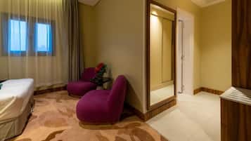 Habitación de hotel con cama, dos sillones morados, una mesita con un florero, un espejo y un armario de madera.