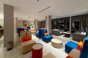 Un vestíbul d'hotel modern amb diverses zones d'assaient, incloent sofàs i butaques, una taula de cafè i un cistell de brossa.