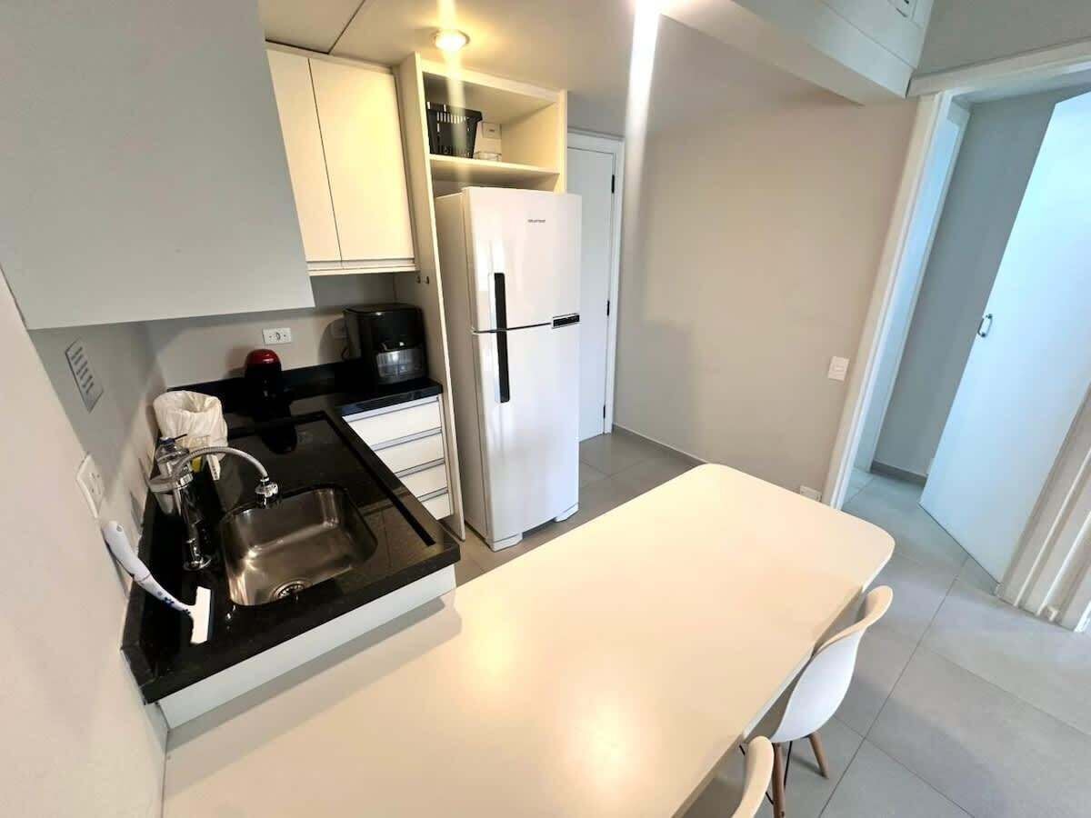 Una cucina compatta con un piano di lavoro bianco, un lavandino in acciaio inossidabile, un frigorifero bianco e un tavolo da pranzo bianco con una sedia.