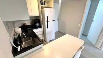 Une cuisine compacte dotée d’un plan de travail blanc, d’un évier en acier inoxydable, d’un réfrigérateur blanc et d’une table à manger blanche avec une chaise.