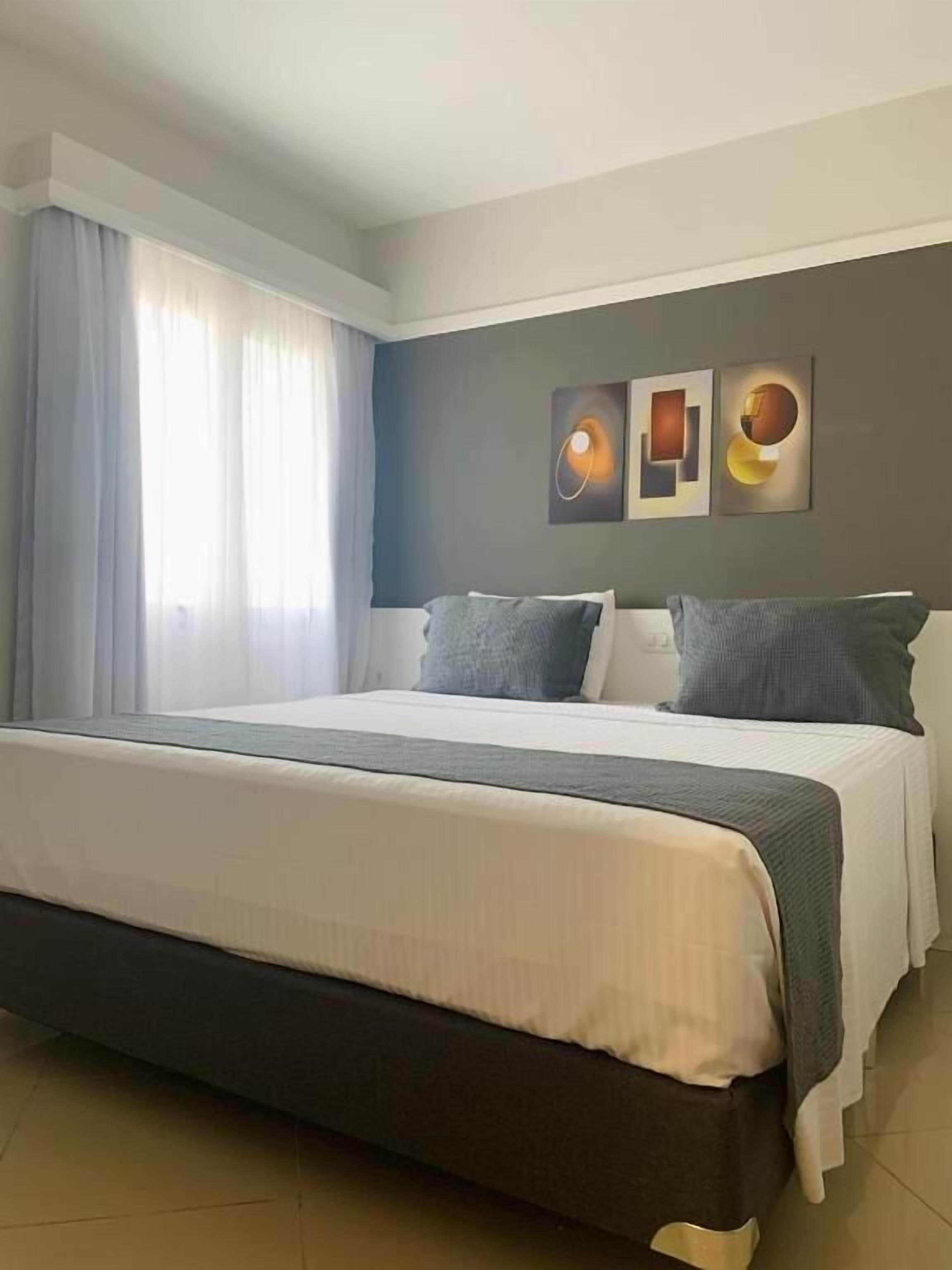 Una camera d'albergo con un letto grande, una finestra con tende e due opere d'arte appese al muro.