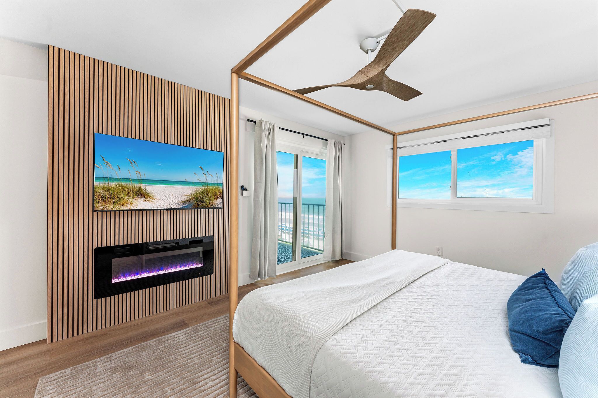 Une chambre avec un lit, un téléviseur fixé au mur, qui est recouvert d’un panneau en bois, un ventilateur de plafond et une fenêtre donnant sur la plage.