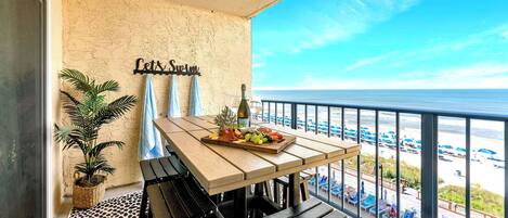 Un balcón con una mesa lista para comer, una planta en maceta y vista a la playa.