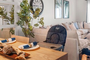 A living room with a sofa, a dining table with plates and a bowl, and a baby stroller. - NEW! Wet Bar, Beachfront, Fully Renovated, 3 Pools, Hot Tub, Gym (Panama City Beach)