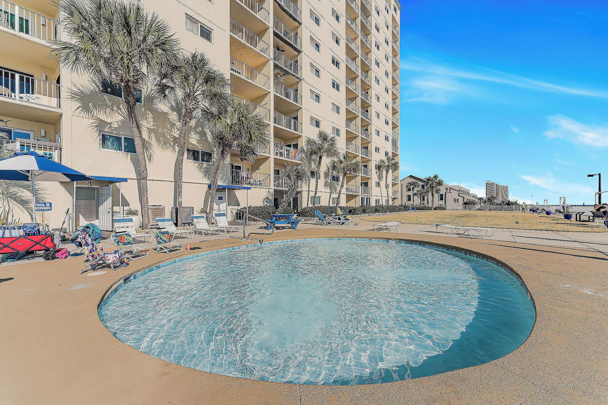 Une piscine entourée de chaises longues et de parasols, elle-même bordée d’un immeuble à plusieurs étages doté de balcons.