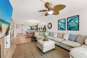 A modern living room with a large sofa, a coffee table, and a ceiling fan. There is a kitchen in the background with a dining area. - NEW! Wet Bar, Beachfront, Fully Renovated, 3 Pools, Hot Tub, Gym (Panama City Beach)