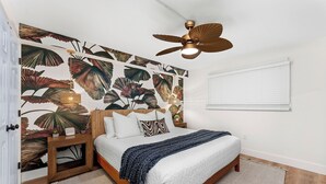 Een slaapkamer met een bed, nachtkastje, plafondventilator, wand met een bladmotief en een raam met jaloezieën.