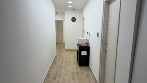 A narrow hallway with a wooden floor, white walls, a clock on the wall, and a door at the end.