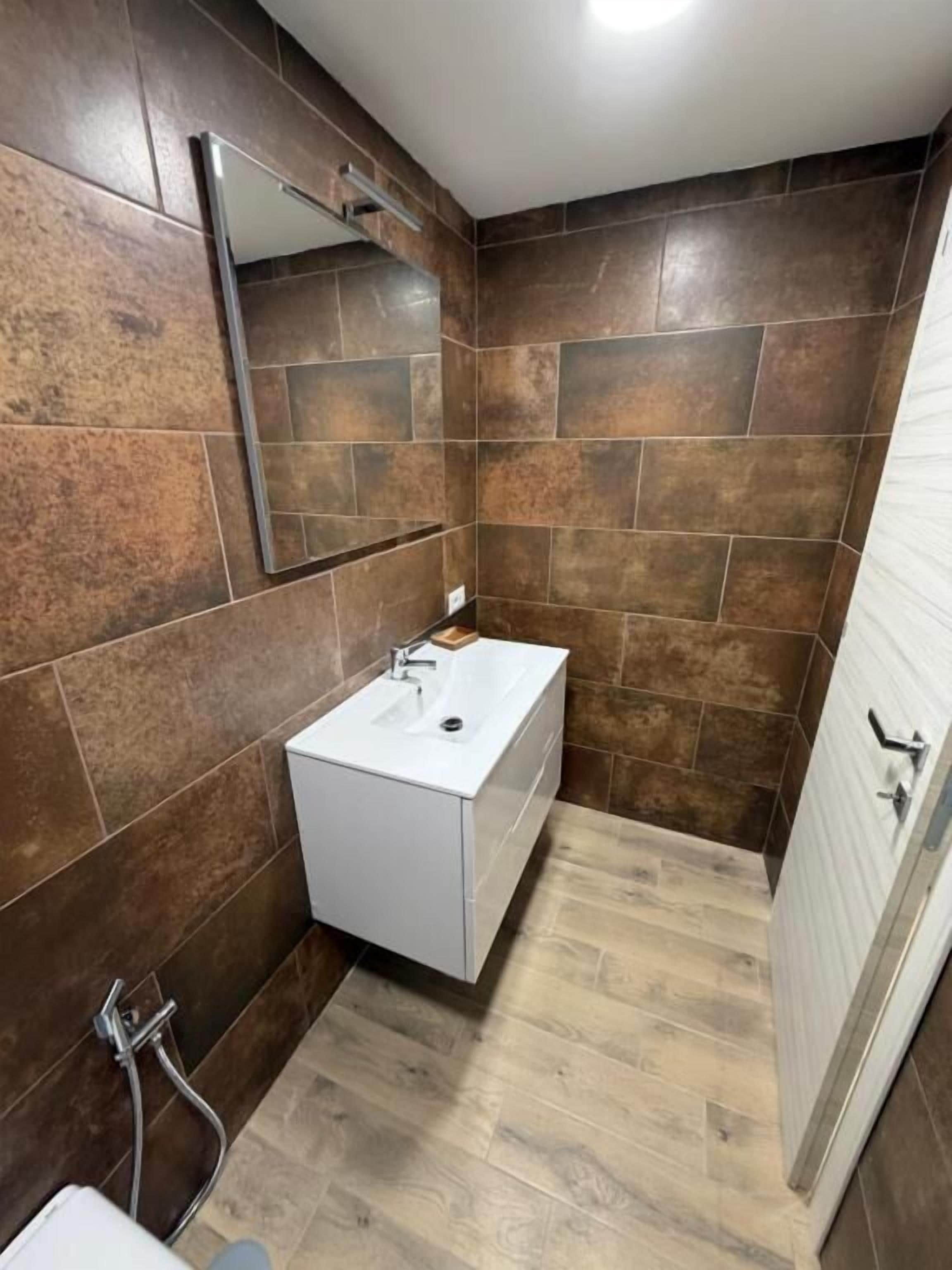 Un baño con lavamanos blanco, espejo y paredes con azulejos marrones.