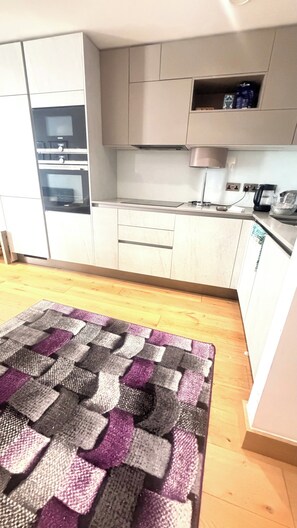 A kitchen with white cabinets, a built-in oven, and a microwave. There is a wooden floor and a patterned rug. - Apartment Sleeps 6 With Balcony (London)