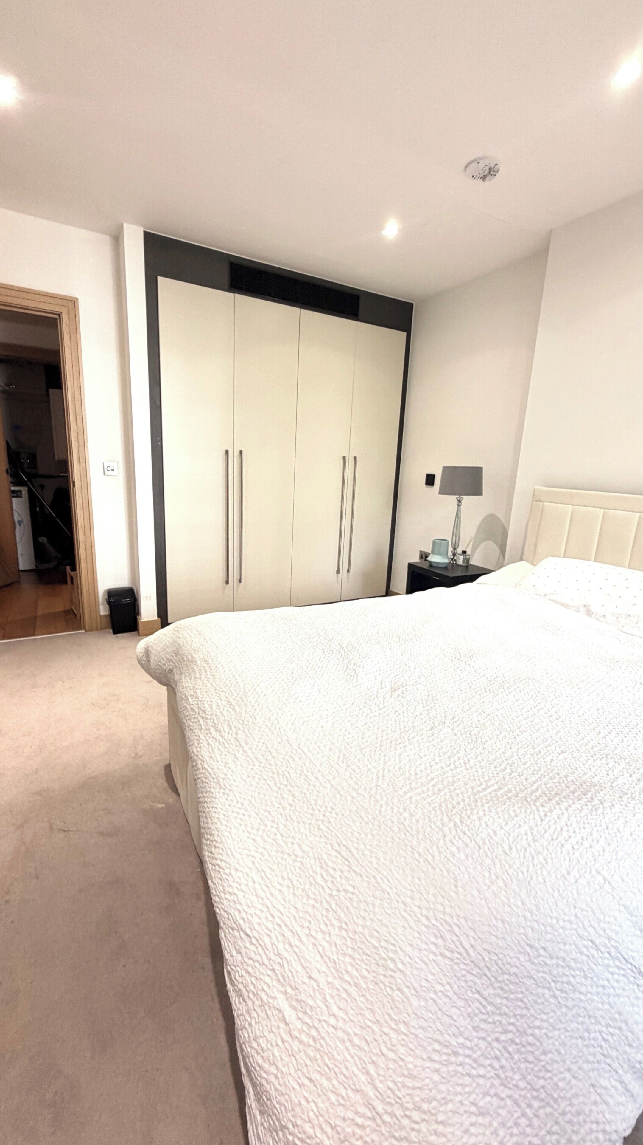 A bedroom with a large bed, a wardrobe, a nightstand with a lamp, and a door leading to a bathroom.