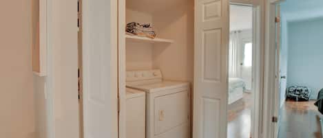 A laundry room with a washing machine and dryer, a shelf with folded clothes, and a door leading to another room.