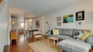 A modern living room with a grey sofa, a dining area with a wooden table, and a kitchen in the background. - Spacious 1,250 sq ft 2-Level 3BR Condo + Parking in Petite-Bourgogne, Montreal (Montréal)