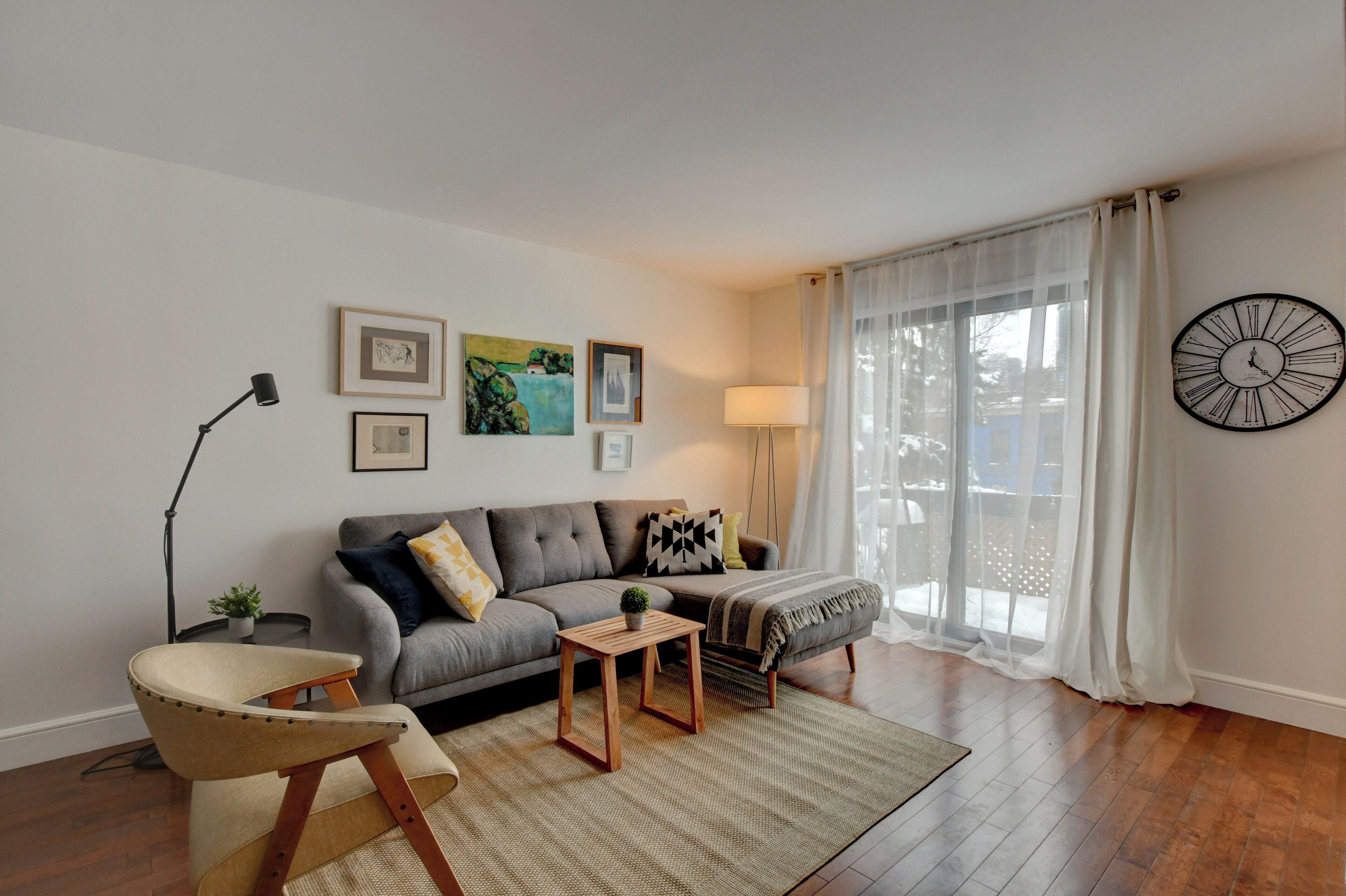 Una sala de estar con sofá, sillón, mesa de centro y una lámpara. Se ven cuadros en la pared y un reloj.