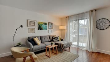 Una sala de estar con sofá, sillón, mesa de centro y una lámpara. Se ven cuadros en la pared y un reloj.