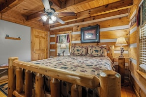A wooden cabin bedroom with a bed, a ceiling fan, a window with curtains, and a wall-mounted picture. - New Listing Hot Tub, Pool Table, Free Attractions (Sevierville)