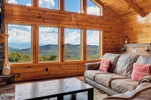 A wooden cabin interior with a leather sofa, a coffee table, and a view of mountains through large windows. - New Listing, Game Room, Hot Tub, Free Attractions, Mountain View (Sevierville)