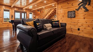 A cozy cabin interior with a wooden ceiling, a leather sofa with gaming-themed pillows, a wooden staircase, and a large window offering a view of the mountains. - New Listing, Game Room, Hot Tub, Free Attractions, Mountain View (Sevierville)