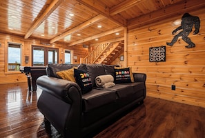 A cozy cabin interior with a wooden ceiling, a leather sofa with gaming-themed pillows, a wooden staircase, and a large window offering a view of the mountains. - New Listing, Game Room, Hot Tub, Free Attractions, Mountain View (Sevierville)