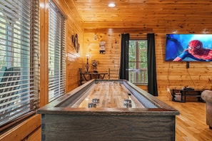 A wooden-floored room with a foosball table, a television displaying a cartoon character, and a window with blinds. - New Listing | 5BR Gatlinburg Cabin w/ Hot Tub + Game Room + Free Tickets (Gatlinburg)