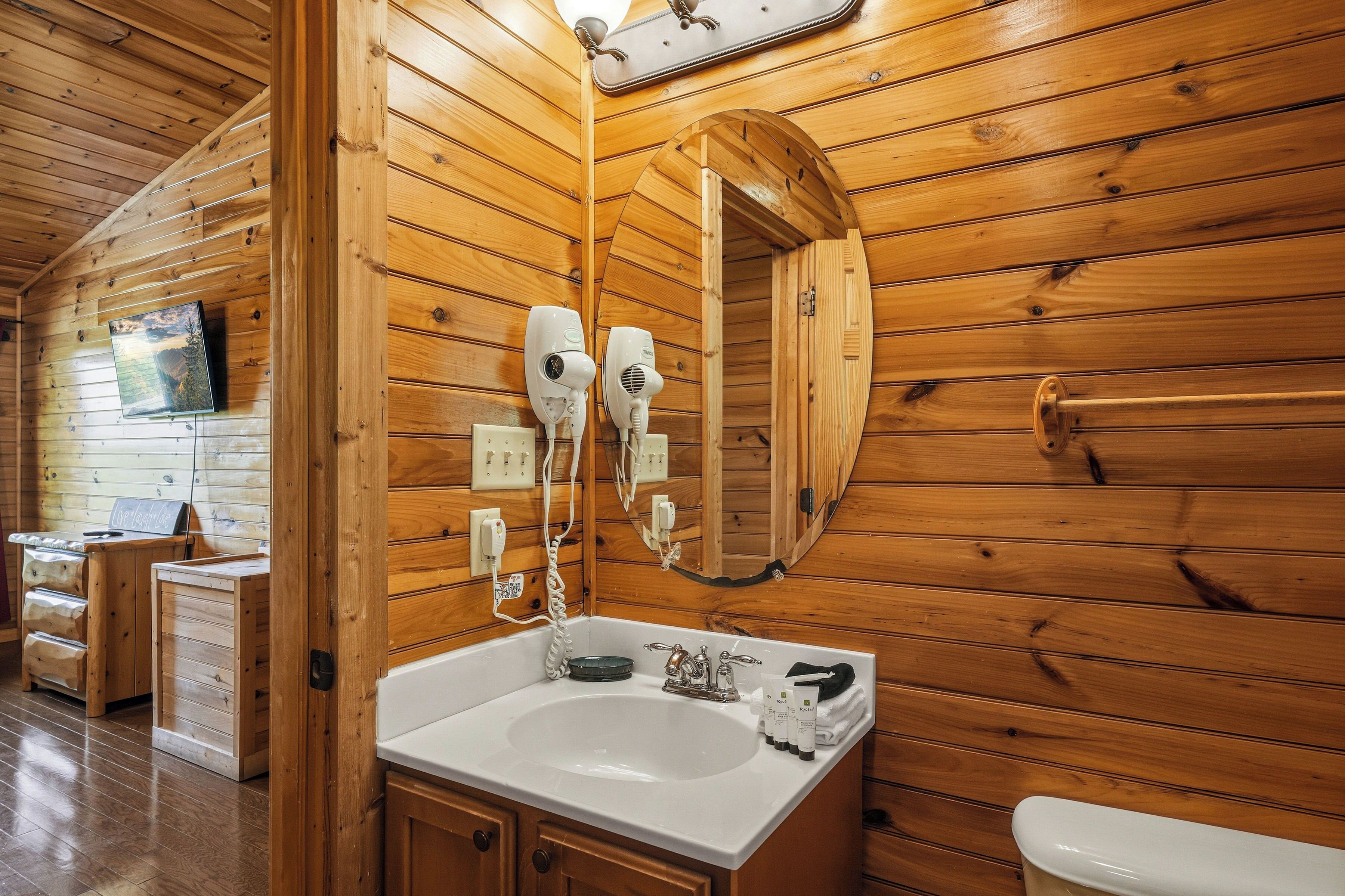 Interior de uma cabana de madeira com uma pia branca, dois secadores de cabelo, um espelho e uma televisão na parede.