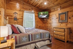 Ein Zimmer mit Holzwänden, einem Bett, einem Fernseher, einer Kommode und einem Fenster mit Jalousien.
