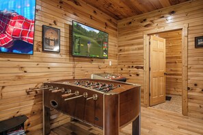 Quarto com piso de madeira, uma mesa de futebol, uma TV montada e um pôster emoldurado de Jurassic Park.