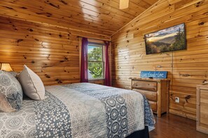 Quarto de cabana de madeira com uma cama, uma janela com cortinas, um quadro na parede e um letreiro com a inscrição 'Live*Laugh*Love'.