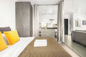 Et moderne hotelværelse med en stor seng, et badeværelse med glasvæg omkring bruseren, og et tekøkken, der ses gennem en åben dør.