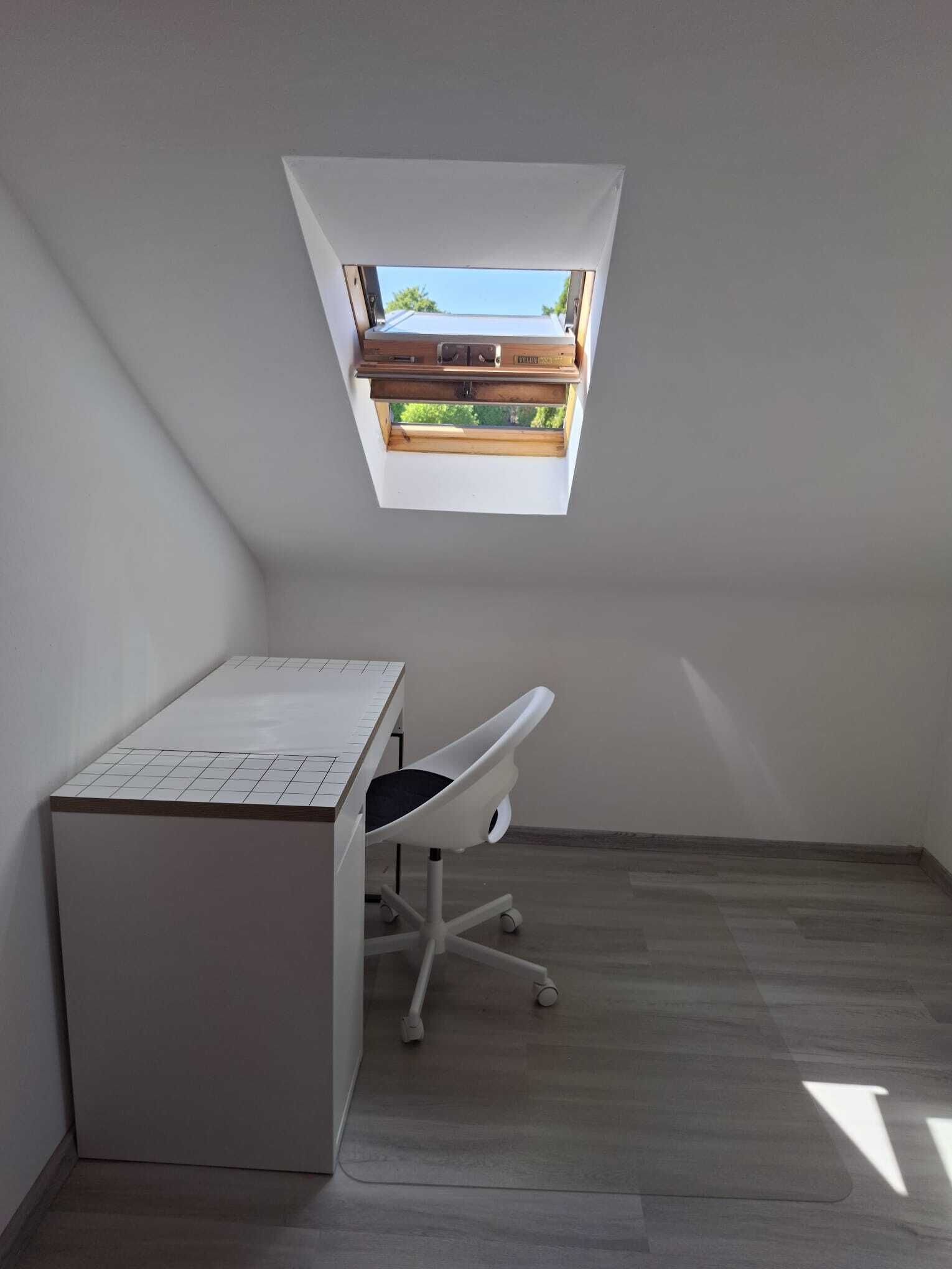 Ein kleiner, moderner Raum mit schrägem Dach, einem Schreibtisch, einem Stuhl und einem Dachfenster.