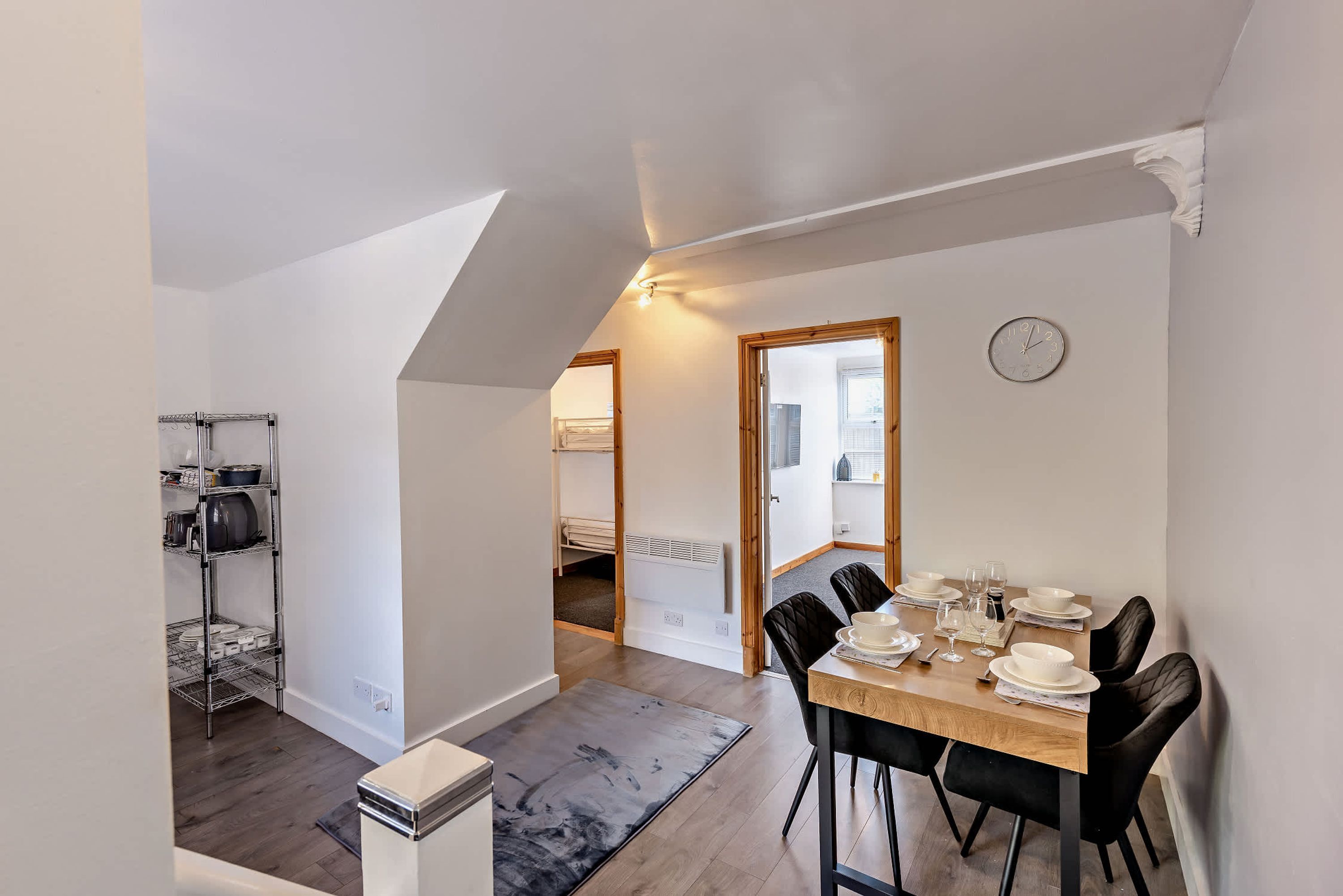 A dining area with a table set for four, a clock on the wall, and a kitchen area with pots and pans on shelves.