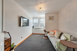 Ein modernes Wohnzimmer mit grauem Teppich, einer grauen Couch mit Kissen, einem Holztisch, einem flachbildfernseher an der Wand und einer Wand mit Ziegeloptik.