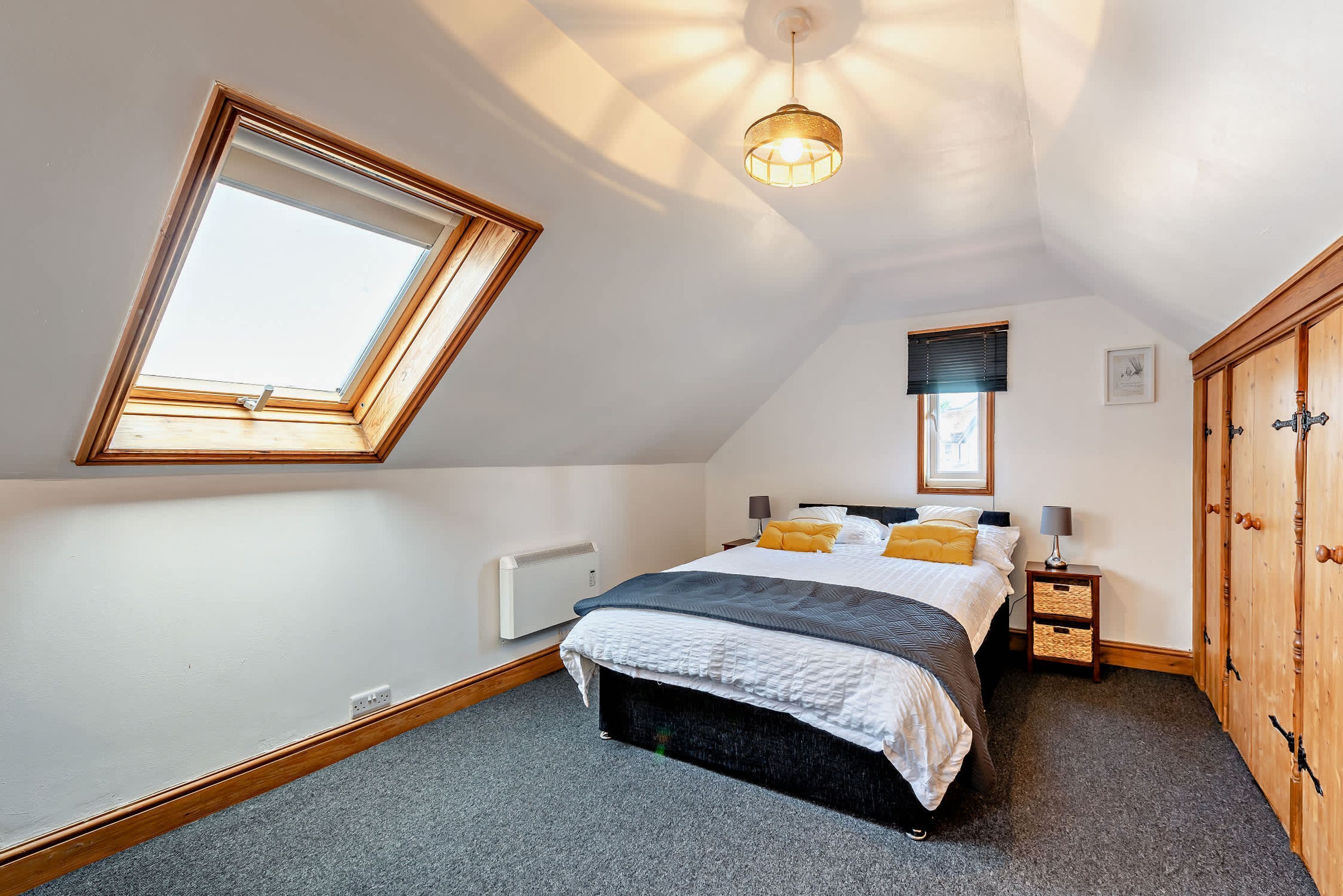 Une chambre avec un puits de lumière, un lit recouvert d’une couette blanche et grise, deux lampes de chevet, une armoire en bois et un radiateur.