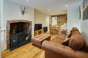 A living room with a brick fireplace, a television, a sofa, and a dining area with a table and chairs. - Host Stay - Hidden Winds Cottage (Guisborough)