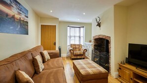 A living room with a brown leather sofa, a plaid ottoman, a fireplace, and a television. - Host Stay - Hidden Winds Cottage (Guisborough)
