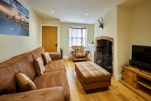 Ein Wohnzimmer mit braunem Ledersofa, einem karierten Hocker, einem Kamin und einem Fernseher.