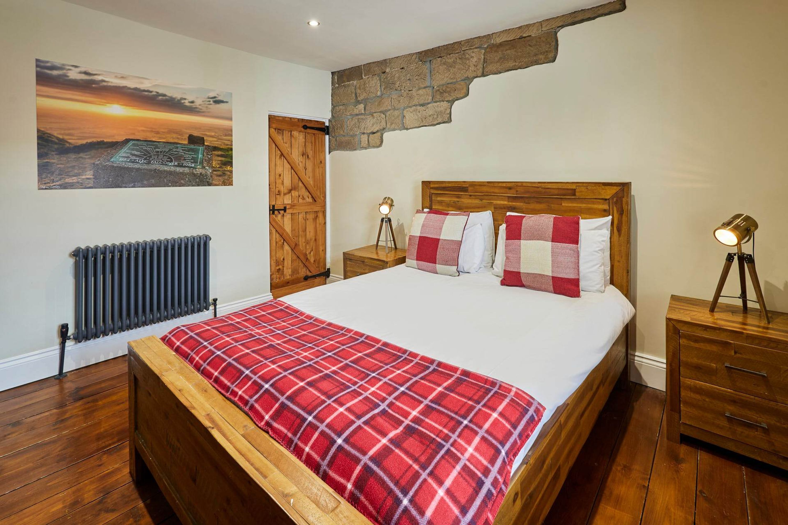 Une chambre à coucher avec un lit, une tête de lit en bois, une couvre-lit à carreaux rouges, une table de chevet avec une lampe, un radiateur et un mur en pierre.