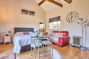 A bedroom with a bed, a red sofa, a table with chairs, a clock, a radiator, and a window with a flowered valance. - Host Stay Snowy Owl Barn (Thetford)