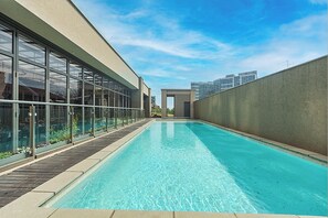 A swimming pool with a clear blue water surface, surrounded by a wooden deck and glass walls. - Sandton Skye apartment, convenience and comfort for your stay (Sandton)