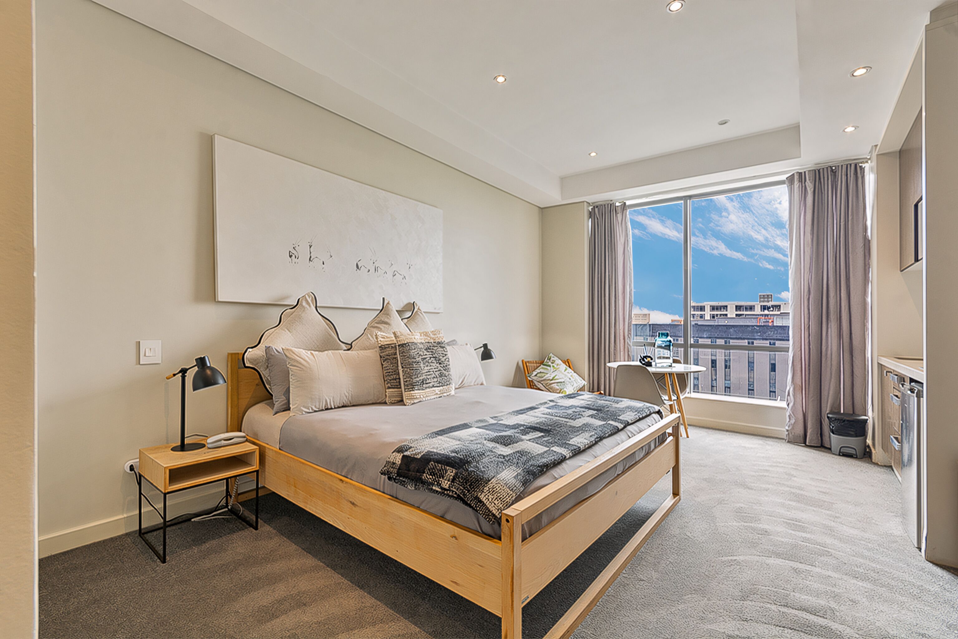 A modern bedroom with a large bed, a bedside table, a lamp, and a view of the city through a large window.