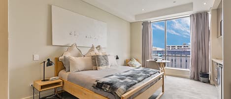 Un dormitorio moderno con una cama grande, una mesita de noche, una lámpara y vistas a la ciudad a través de un amplio ventanal.