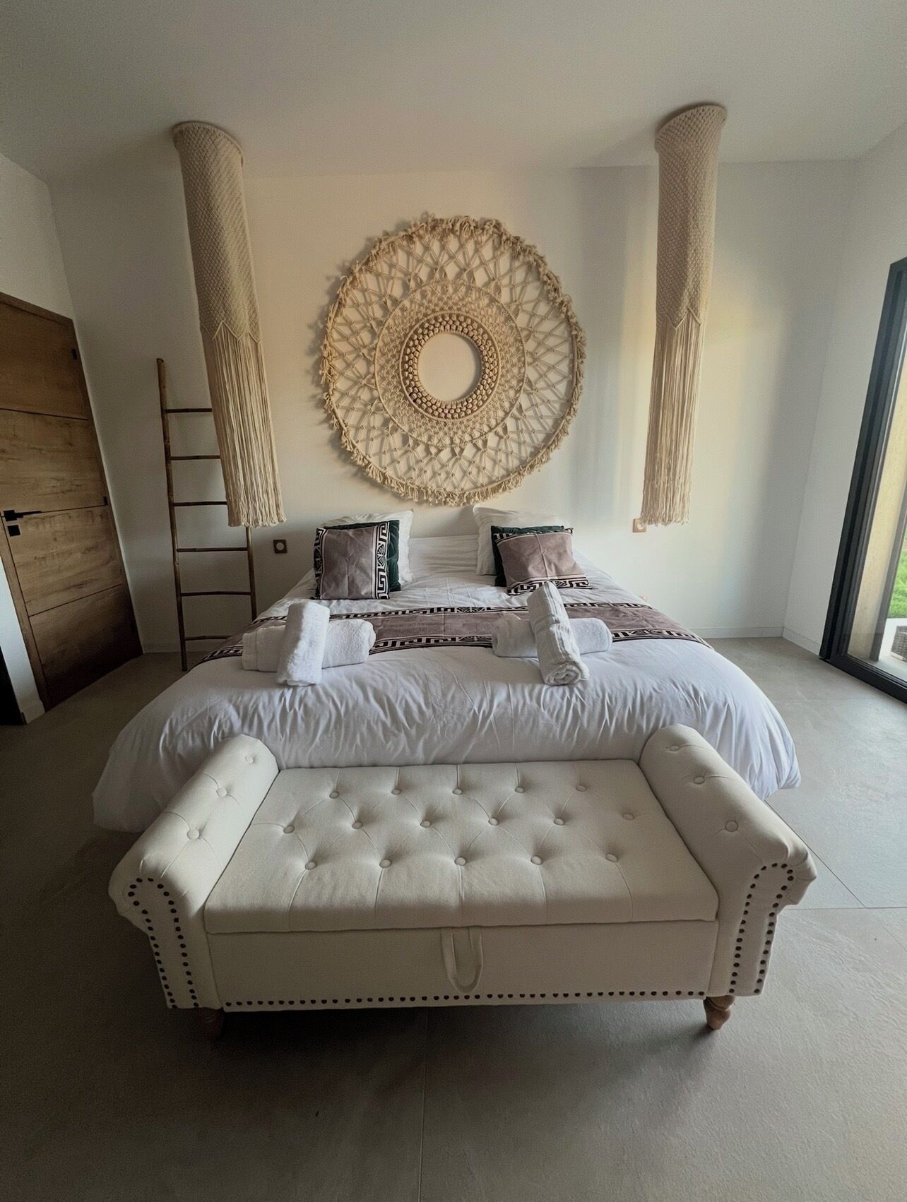 A large bed with a tufted headboard, a macramé wall hanging, and a view of the outdoors.