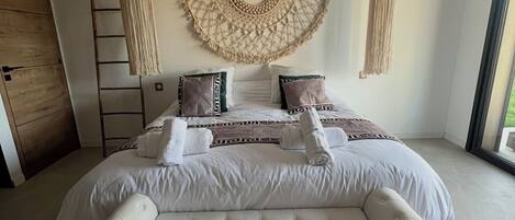 A large bed with a tufted headboard, a macramé wall hanging, and a view of the outdoors.