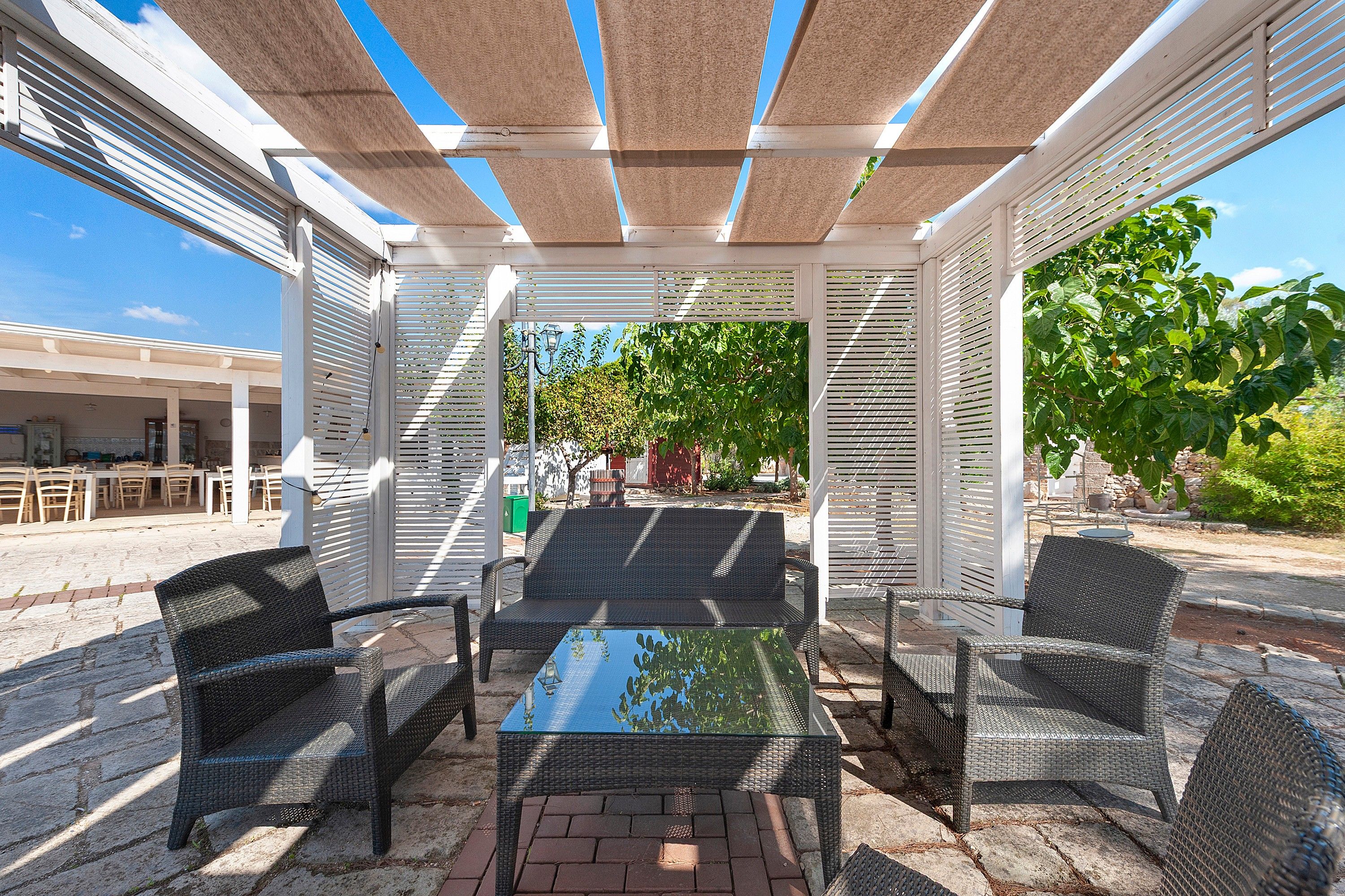 Ein Außenbereich mit einer Terrasse, einem Tisch aus Glas, Korbstühlen und einer Pergola.